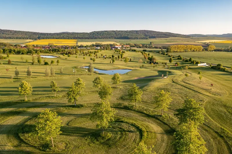 Die Anlage aus der Luft im Abendlicht mit Teich, Fairways und dem Dorf im Hintergrund.