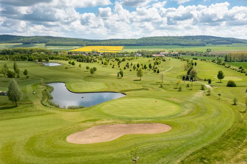 Blick über ein Grün mit vorgelagertem Bunker und Teich, dahinter der Platz und die Hügel.