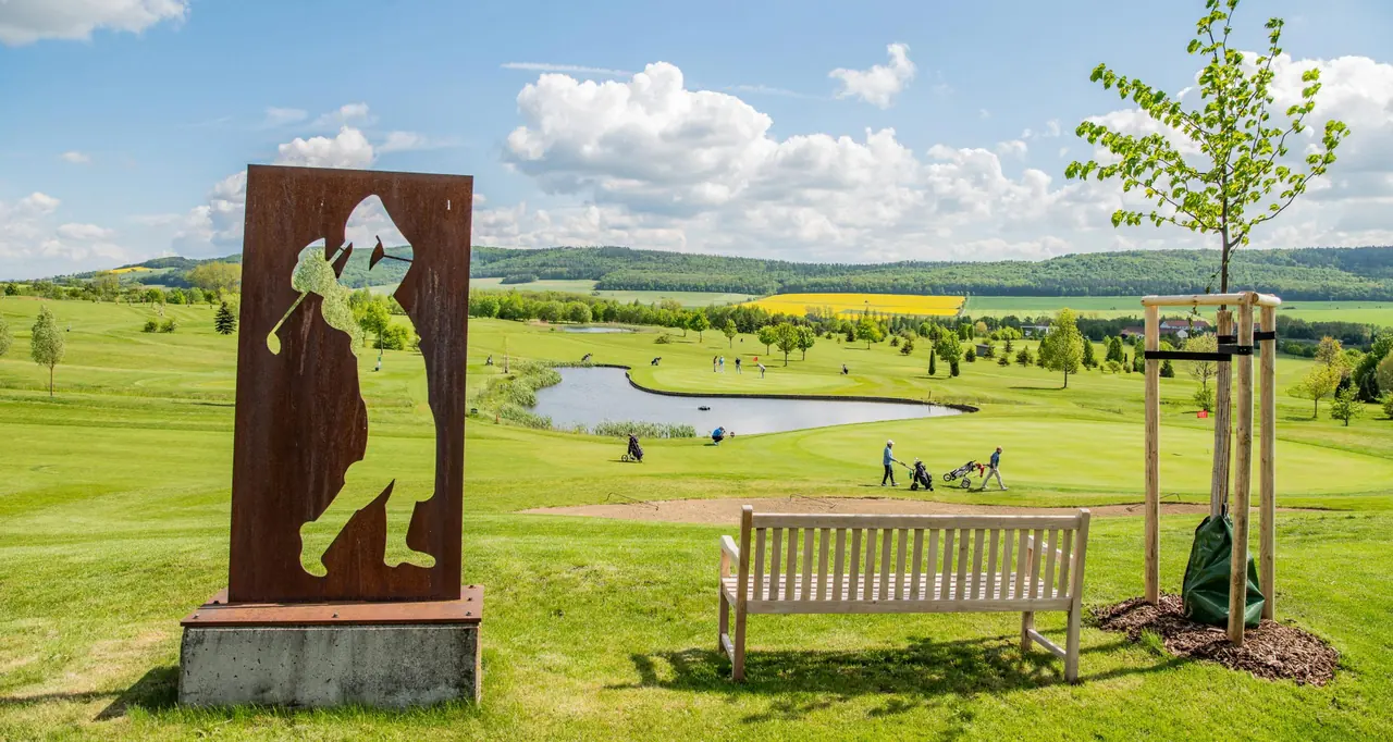 Die Corten-Stahl-Skulptur eines Golfers am ersten Abschlag, dahinter der Platz mit Teich und die Hügel des Hainich.